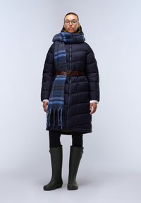 Cappotto imbottito blu navy con cintura, abbinato a una sciarpa a quadretti blu, leggings neri e stivali di gomma verde al ginocchio.