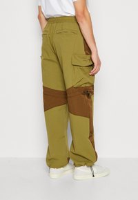 Mand med olivengrønne og brune lapper cargo-bukser med elastisk talje og manchetter, parret med hvide sneakers og en hvid skjorte.