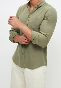 Camisa de botones verde oliva con una tela texturizada, mangas largas arremangadas y un bolsillo en el pecho. Combinada con pantalones blancos.