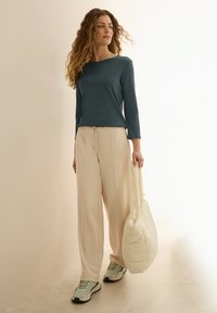 Haut à manches longues couleur sarcelle, pantalon large beige et sac fourre-tout matelassé beige. La mannequin porte des baskets bleu clair. Textures douces avec un design minimal.