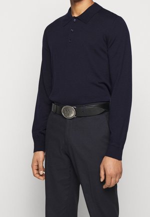 Homme portant un polo bleu marine à manches longues rentré dans un pantalon noir avec une ceinture noire décorée d'une boucle argentée avec un emblème animal.
