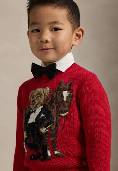 Jonge jongen draagt een rode trui met een ontwerp van een teddybeer en een paard, een wit overhemd met kraag en een zwarte strik, kijkt zelfverzekerd naar de camera.