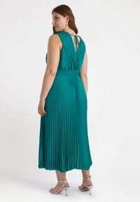 Abito pieghettato in teal con vita aderente e dettaglio della schiena scoperta, caratterizzato da una chiusura con lacci. Il tessuto testurizzato arriva fino al polpaccio. Sandali argentati con tacco.