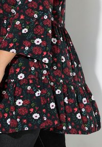 Schwarzes Blumenshirt mit Peplum-Schnitt, das rote und weiße Blumen sowie grüne Blätter zeigt, und einem gestuften Rüschensaum. Weicher Stoff mit fließendem Design.