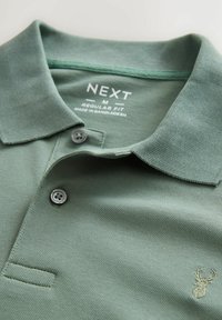 Polo de color verde oliva con tejido texturizado, dos botones y un logo de ciervo bordado en el pecho. La etiqueta indica tamaño M, corte regular.
