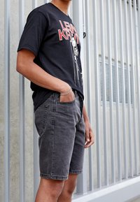 Persona vestida con una camiseta gráfica negra y pantalones cortos de mezclilla grises se encuentra de pie con una mano en el bolsillo, contra un fondo de barandilla de metal.