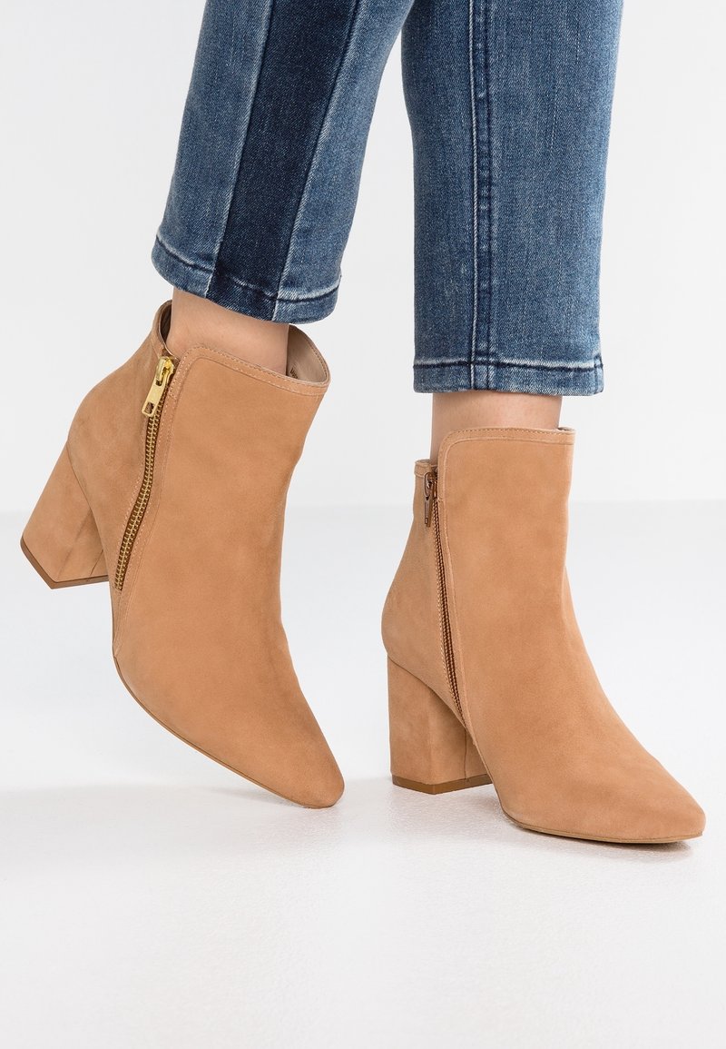 Bottes cheville en daim tan avec talons carrés et fermetures éclair, portées avec un jean en denim bleu sur un fond blanc uni.