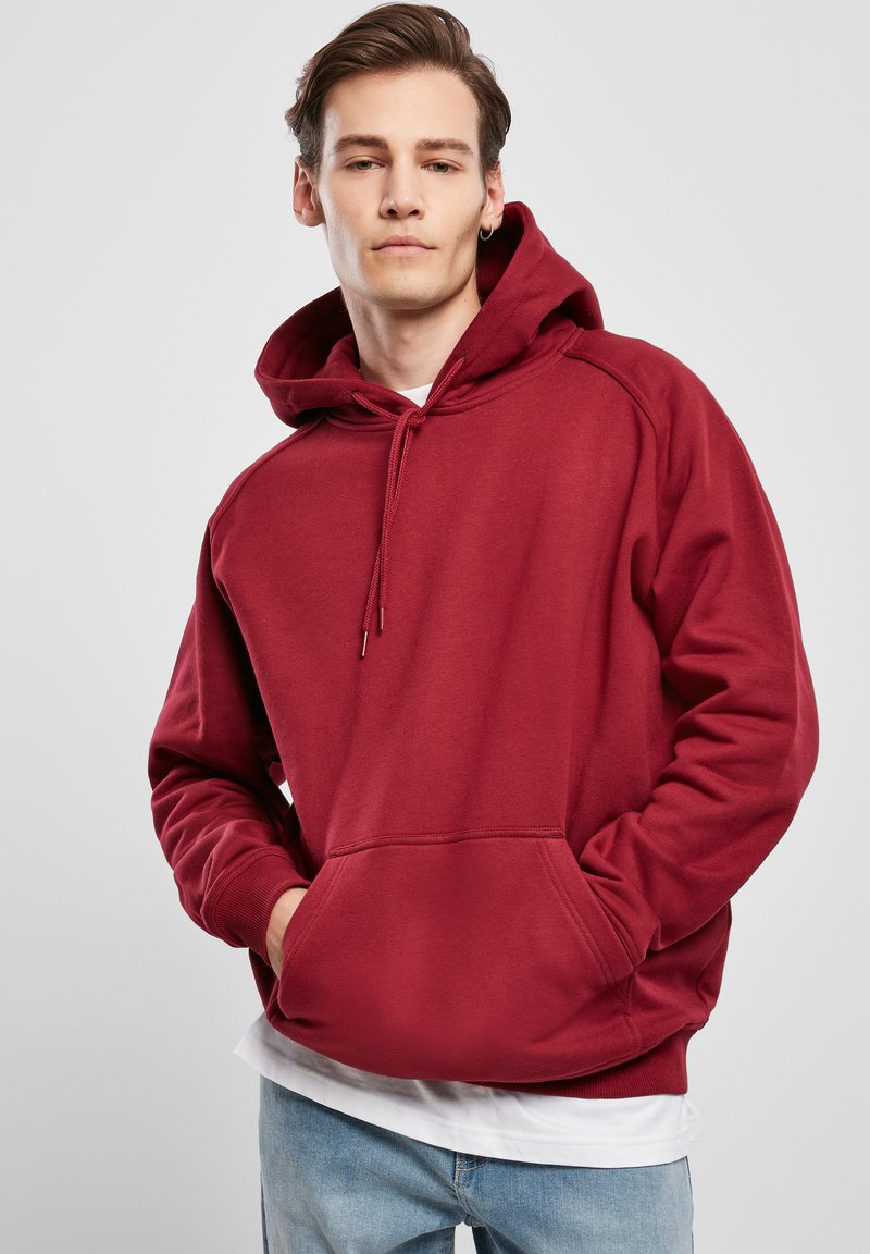 Felpa bordeaux con tasca frontale, cappuccio con cordino, polsini e orlo a coste. Tessuto liscio, vestibilità comoda, indossata sopra una maglietta bianca.