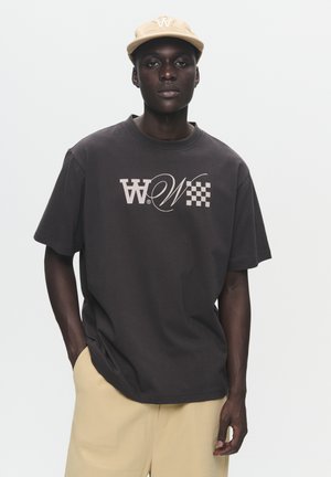 Dark gray oversized t-shirt with a light logo design and checker pattern, paired with beige shorts. The model wears a beige cap.