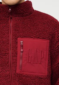 Κοντινό πλάνο κόκκινου φλις μπουφάν με φερμουάρ και μεγάλη τσέπη στο στήθος με ανάγλυφο λογότυπο "GAP".