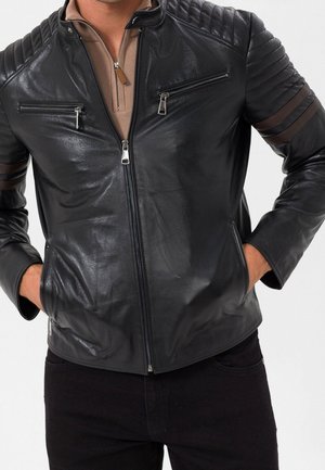 Uomo che indossa una giacca di pelle nera con strisce marroni sulle maniche e tasche con zip, mani nelle tasche laterali, sopra una camicia marrone con zip.