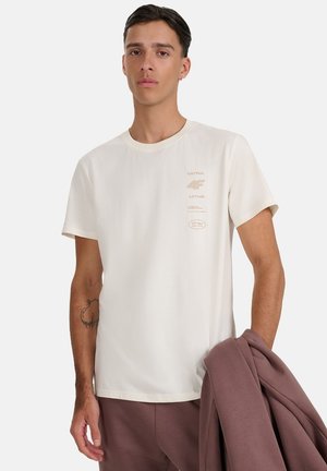 Jasnobeżowa bawełniana koszulka z krótkimi rękawami, okrągłym dekoltem i nadrukowanymi akcentami logo na piersi. Model trzyma brązową kurtkę.