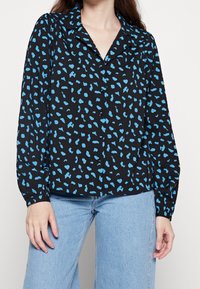 Blusa nera con stampa leopardata blu, maniche lunghe, design con colletto, tessuto leggero, chiusura a bottoni e vestibilità rilassata. Abbinata a jeans azzurri.