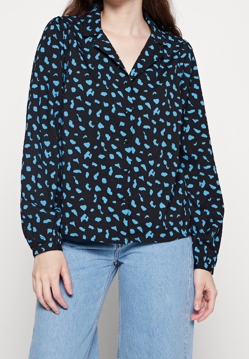 Blusa nera con stampa leopardata blu, maniche lunghe, design con colletto, tessuto leggero, chiusura a bottoni e vestibilità rilassata. Abbinata a jeans azzurri.