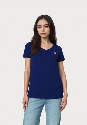 Nuori nainen, jolla on pitkät tummat hiukset, pukeutuneena laivaston siniseen V-kaulaiseen t-paitaan ja vaaleansinisiin farkkuihin, seisomassa yksinkertaisen valkoisen taustan edessä.