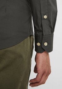 Donkergroen overhemd met knopen en een gladde textuur, voorzien van lichtgekleurde knopen op de mouwen. Gekleed met olijfgroene broek.