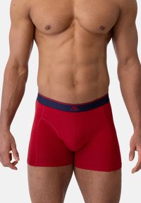 Červené boxerky s tmavě modr�ým pasem, které mají těsný střih a hladkou texturu. Design obsahuje střední kapsu pro oporu.