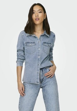 Cămașă din denim de culoare albastru deschis, cu deschidere cu nasturi în față, două buzunare pe piept și o guler clasic. Prezintă mâneci răsucite și o croială lejeră.