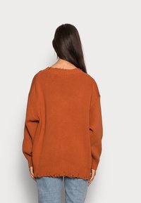 Rostfärgad oversized tröja med ribbad halsringning och fransig fåll. Mjuk stickad textur och sänkta axlar. Matchad med ljusblå denim.