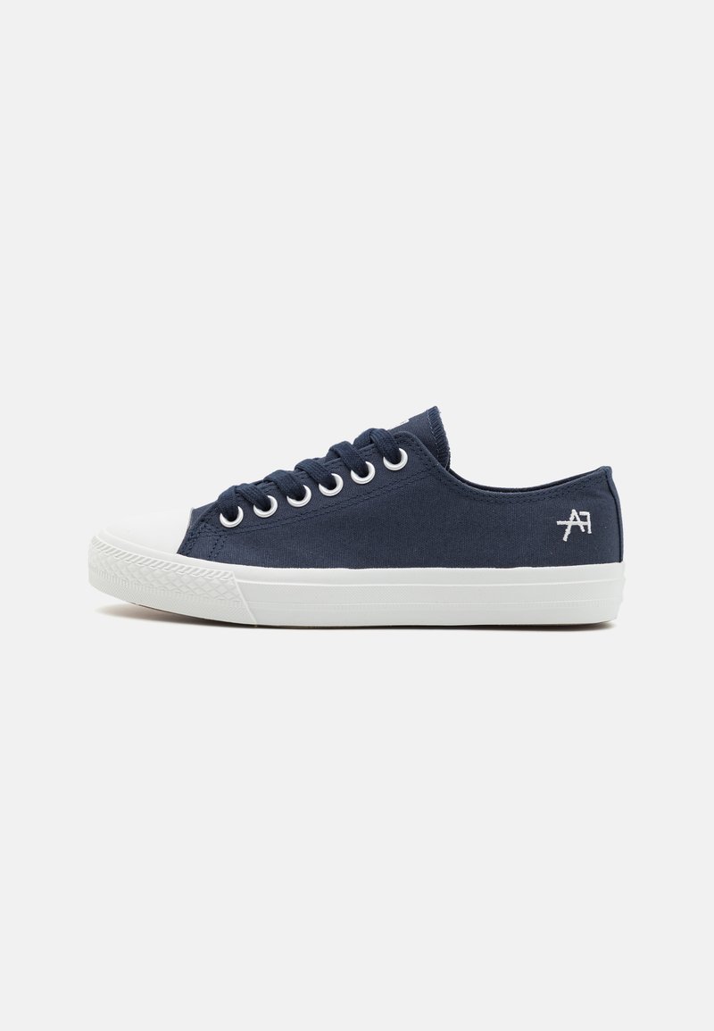 Marineblå lerret sneakers med hvit gummikappe og såle. Har avrundet form, snøring og diskret logo detalj på siden.