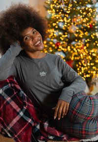 Grijs shirt met lange mouwen met de tekst "Jolly", gecombineerd met een geruit deken in rood, marineblauw en wit. Op de achtergrond staat een zacht verlichte kerstboom.