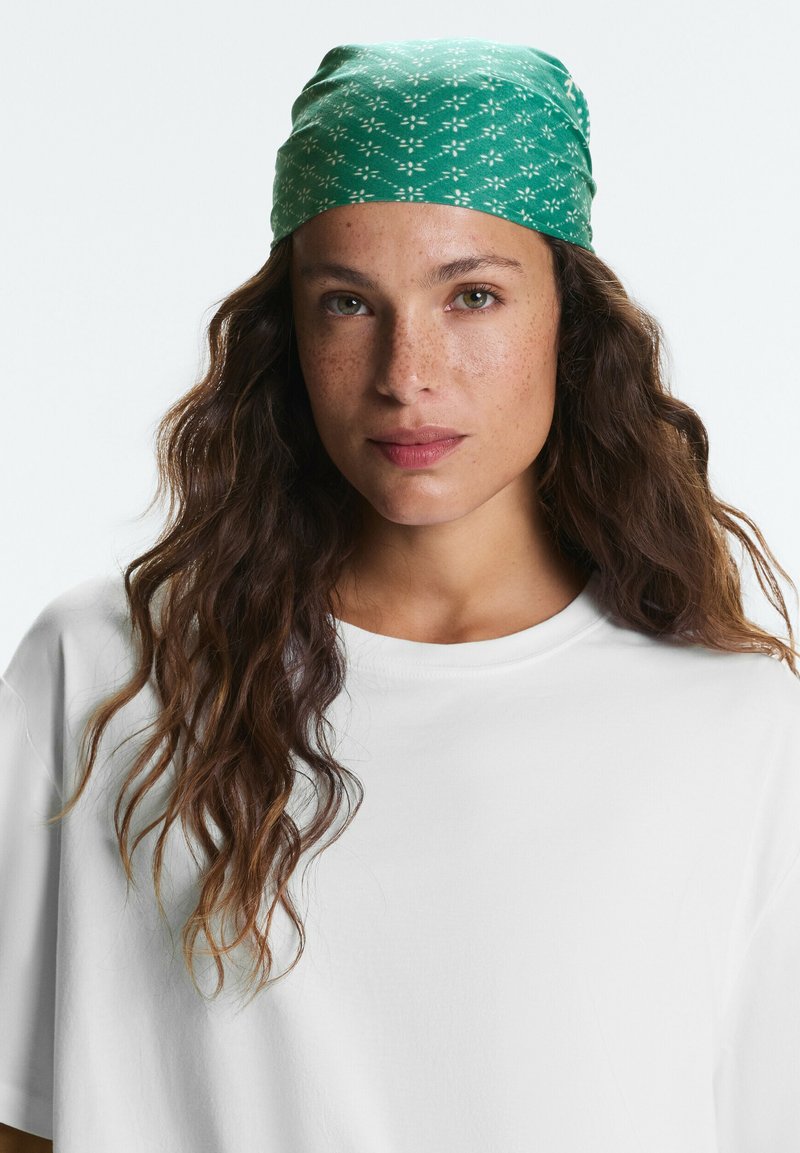 Femme aux longs cheveux bruns ondulés portant un foulard vert à motifs et un t-shirt blanc uni, regardant directement la caméra.