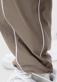 Pantaloni sportivi marroni con dettagli in piping bianco, realizzati in tessuto leggero, dotati di una vita elastica e polsini arricciati, abbinati a scarpe bianche.