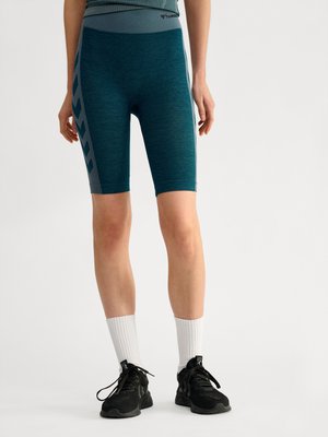 Onderlichaam van een persoon die turquoise fietsbroek draagt met zijstrepen, witte geribde sokken en zwarte sportschoenen op een witte achtergrond.
