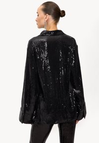 Veste-chemise noire à sequins avec col, présentant une surface texturée et brillante ainsi qu'une coupe ample. Manches longues se terminant par de larges poignets.