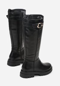 Bottes en cuir noir montantes avec une semelle en caoutchouc robuste, dotées d'une fermeture éclair sur le côté et d'une sangle ajustable avec une boucle dorée.