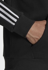 Svart hoodie med kontrasterande vita ränder på ärmarna, texturerad tygkvalitet och en framsida ficka utformad för tillgänglighet.