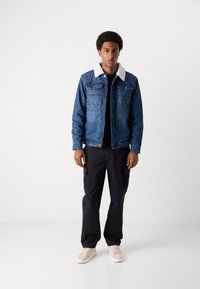 Jeansjacke in Blau mit einem weißen Kunstfellkragen, ausgestattet mit Brusttaschen und einer vordernen Knopfleiste, getragen über einem schwarzen Hemd.