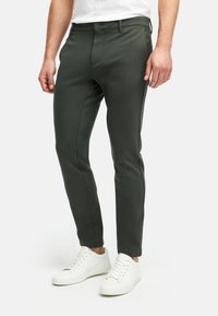 Pantalones ajustados de color verde oscuro con una textura suave, con cierre de botón y trabillas para cinturón, combinados con zapatillas blancas.
