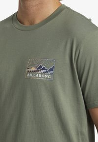 Olive green cotton t-shirt featuring a printed logo in a rectangular design with sun and mountain graphics. Short sleeves and crew neckline.