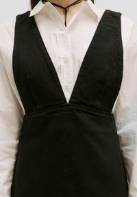 Zwarte corduroy jurk met een diepe V-halslijn, voorzien van een aansluitend lijfje en brede schouderbanden, gedragen over een witte blouse met kraag.