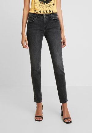 Kobieta ubrana w czarne, opinające, z wysokim stanem dżinsy, żółtą bezrękawnikową bluzkę z nadrukiem oraz czarne sandały na obcasie z odkrytymi palcami na jednolitym tle.