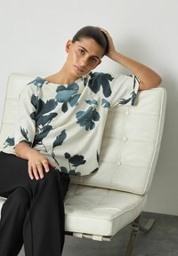 Blus med blommönster i vitt och mörkblått, med en lös passform och korta ärmar, i kombination med svarta byxor, sittande.