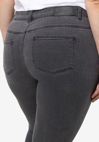 Primer plano de una persona que lleva jeans oscuros ajustados color gris con bolsillos traseros y una etiqueta negra en la cinturilla, con la mano reposando a un costado.