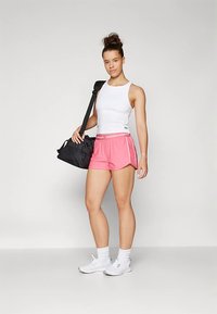 Czerwone sportowe spodenki z białymi akcentami, zestawione z białym tank topem i białymi butami sportowymi. Na ramieniu zawieszona jest czarna torba typu duffel.