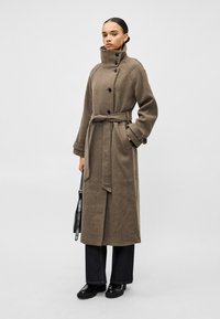 Casaco trench coat de lã castanha com gola alta, fecho de botões e cinto. Apresenta bolsos laterais e punhos largos. Usado com calças de ganga escuras e sapatos.