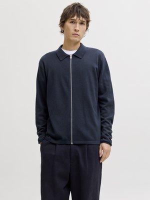 Noor mees lühikeste pruunide juustega, kandes tume-sinist lukuga kampsunit valge särgi kohal, seisab käed taskutes ühevärvilise tausta ees.