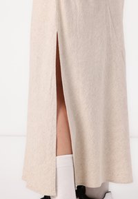 Gonna lunga beige con uno spacco laterale, realizzata in tessuto testurizzato. L'orlo è leggermente curvo, rivelando un calzino bianco e una scarpa nera.