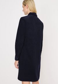 Femme blonde portant une robe noire marine à manches longues, arrivant au genou avec un col, vue de dos sur un fond blanc.