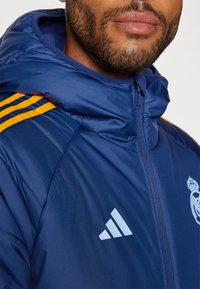 Giacca imbottita blu navy con cappuccio, dotata di dettagli a righe arancioni, logo Adidas bianco e scudetto del Real Madrid sul petto.