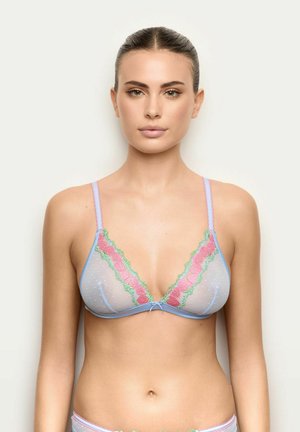 Mujer con el pelo peinado hacia atrás, vistiendo un sujetador de tul azul claro con detalles de encaje en rosa y verde, de pie contra un fondo liso y claro.