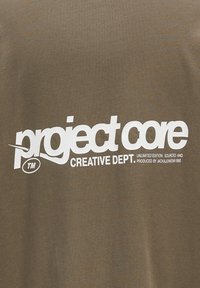 Camiseta marrón con gran texto blanco que dice "project core CREATIVE DEPT." y detalles adicionales. Fabricada en algodón con textura suave.
