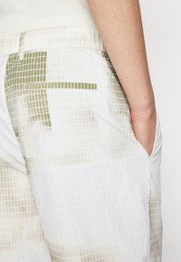 White fabric shorts featuring a grid pattern in green and beige, with side pockets, and a Calvin Klein label on the back pocket.