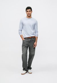 Lichtblauw overhemd met lange mouwen, grijze spijkerbroek, bruine leren riem, witte sneakers met bruine zolen, staand tegen een effen achtergrond.