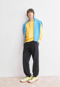 Gele sporttop met rits, blauwe sweatshirt over de schouders gehangen, zwarte joggingsbroek en gele sneakers met rode accenten. Binnenomgeving.