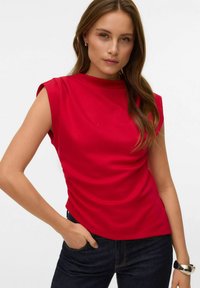 Top rojo de manga corta con cuello alto y redondeado y un diseño ajustado. La tela parece lisa, complementando unos vaqueros oscuros.